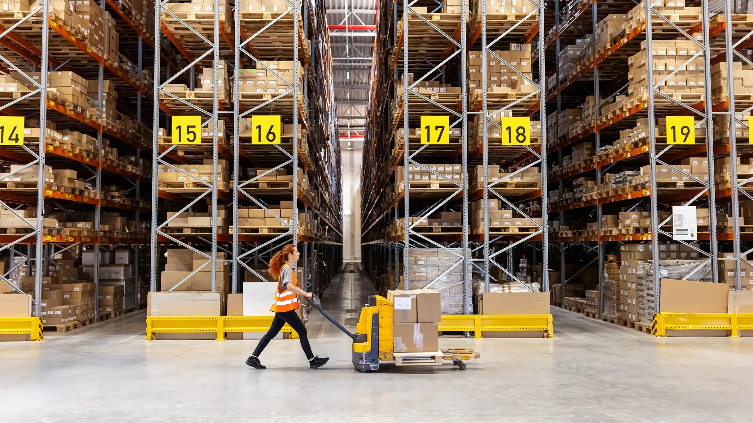 A warehouse worker wearing a safety vest pulls a loaded manual pallet jack down an aisle of high-density storage racks.