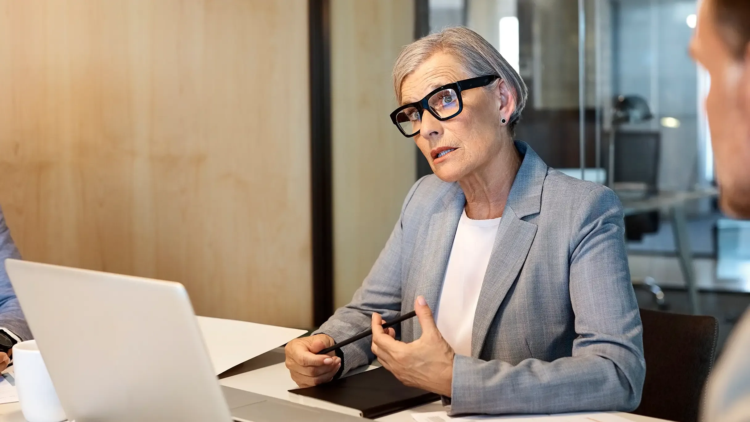 A business woman wearing thick glasses and a grey blazer gestures while speaking in an office.