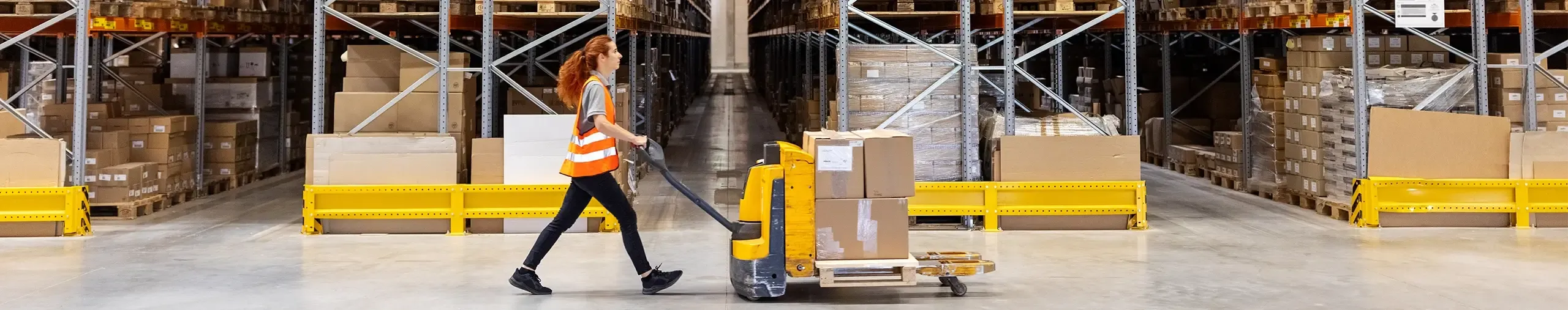 A warehouse worker wearing a safety vest pulls a loaded manual pallet jack down an aisle of high-density storage racks.